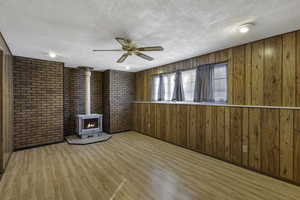 Unfurnished living room featuring wood finished floors, a wood stove, a ceiling fan, a textured ceiling, and wooden walls