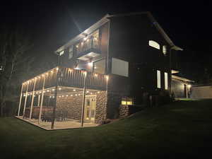Back of house at night featuring a lawn, a patio area, a balcony, and stone siding