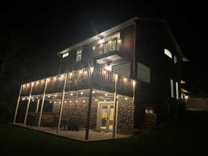 Back of house at night featuring a patio, stone siding, a yard, and a balcony