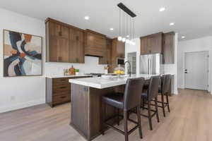 Enhanced staged photo Kitchen with a breakfast bar area, stainless steel appliances, an island with sink, light wood finished floors, and hanging light fixtures