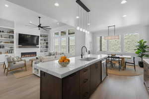 Enhanced staged photo Kitchen featuring dark wood finish cabinetry, decorative light fixtures, an island with sink, a fireplace, and light stone countertops