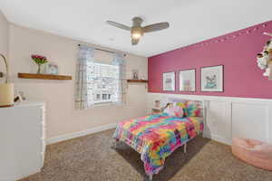 Carpeted bedroom featuring ceiling fan, wainscoting, and a decorative wall