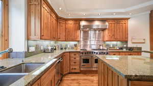 Kitchen with range with two ovens, wall chimney range hood, recessed lighting, light stone counters, and light wood-style floors