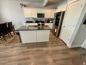 Kitchen with stainless steel appliances, white cabinetry, dark stone countertops, a kitchen island with sink, and beam ceiling