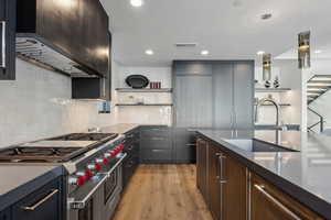 Kitchen with open shelves, range with two ovens, wall chimney exhaust hood, light wood finished floors, and hanging light fixtures