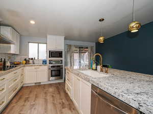 Kitchen with appliances with stainless steel finishes, white cabinets, hanging light fixtures, light wood finished floors, and light stone counters