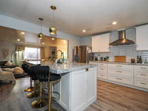 Kitchen with white cabinetry, open floor plan, light wood-style flooring, a breakfast bar area, and wall chimney range hood