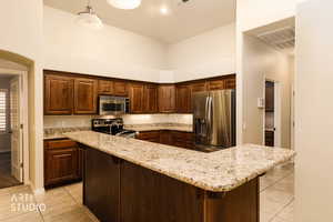 Kitchen with stainless steel appliances, a kitchen breakfast bar, light stone countertops, a high ceiling, and hanging light fixtures