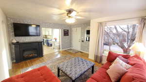 Living room with light wood-type flooring, a glass covered fireplace, ceiling fan, hand painted walls, and a textured ceiling