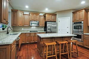 Kitchen featuring a kitchen breakfast bar, light stone countertops, stainless steel appliances, dark wood-style flooring, and brown cabinets