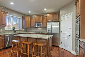 Kitchen with brown cabinets, a breakfast bar area, appliances with stainless steel finishes, light stone countertops, and recessed lighting