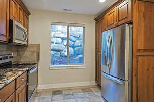 Kitchen featuring appliances with stainless steel finishes, brown cabinets, dark stone counters, stone tile flooring, and decorative backsplash
