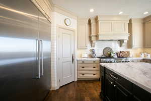 Kitchen with cream cabinets, dark cabinetry on the island, stainless steel built in refrigerator, Calcutta marble counters, and dark wood finished floors