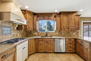 Kitchen with custom range hood, brown cabinets, stainless steel appliances, light stone countertops, recessed lighting, gas stove and double ovens