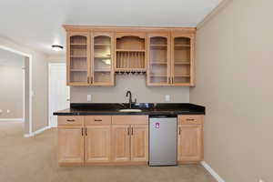 Wet bar featuring dark countertops, crown molding, stainless steel dishwasher, light carpet, and light brown cabinetry