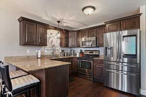 Kitchen with stainless steel appliances, dark brown cabinets, a peninsula, and light stone countertops