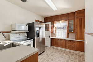 Kitchen featuring brown cabinetry, white appliances, light countertops, glass insert cabinets, and light flooring