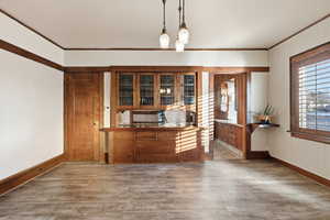 Bar with decorative light fixtures, wood finished floors, ornamental molding, brown cabinets, and glass insert cabinets