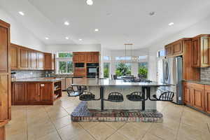 Kitchen featuring backsplash, brown cabinetry, a kitchen bar, a kitchen island with sink, and recessed lighting