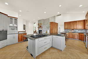 Two tone kitchen featuring a large fireplace, stainless steel oven, dark stone counters, light tile patterned floors, and vaulted ceiling
