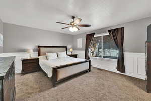 Bedroom featuring a wainscoted wall, a decorative wall, light carpet, ceiling fan, and a textured ceiling
