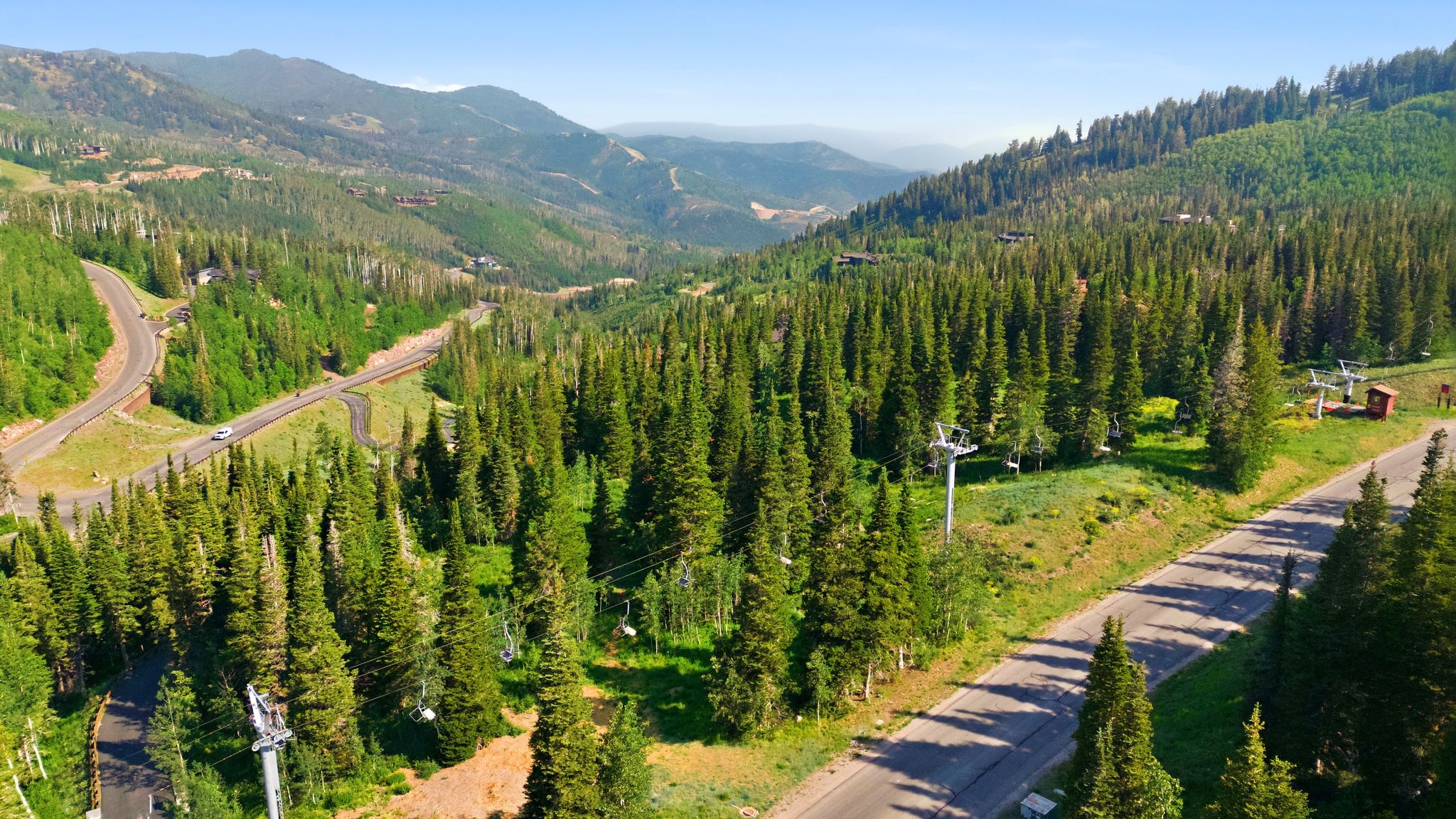 Mountain landscape featuring extensive evergreen forests, winding paved roads, distant peaks, and ski lift infrastructure
