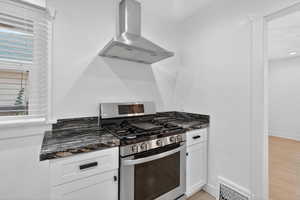 Kitchen with stainless steel gas range, wall chimney exhaust hood, white cabinetry, dark stone countertops, and light wood-type flooring