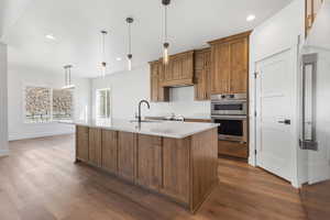 Kitchen featuring brown cabinetry, decorative light fixtures, stainless steel appliances, a kitchen island with sink, and dark wood-style flooring