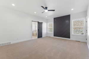 Unfurnished bedroom featuring a barn door, ensuite bath, light carpet, a ceiling fan, and recessed lighting