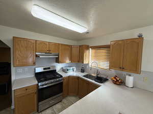 Kitchen with stainless steel appliances, light countertops, a textured ceiling, brown cabinetry, and under cabinet range hood
