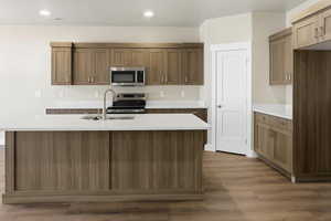 Kitchen with stainless steel appliances, a center island with sink, brown cabinets, dark wood-type flooring, and recessed lighting