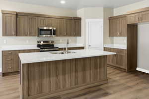 Kitchen with appliances with stainless steel finishes, light wood finished floors, an island with sink, light stone counters, and brown cabinets