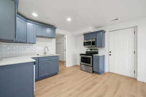 Kitchen with appliances with stainless steel finishes, blue cabinetry, backsplash, light wood-style flooring, and light stone countertops