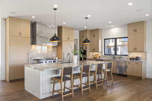 Main Kitchen with light brown cabinetry, light stone counters, decorative light fixtures, dark wood-style flooring, and an island with sink