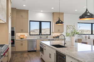 Kitchen with light brown cabinetry, light stone countertops, hanging light fixtures, beverage cooler, and appliances with stainless steel finishes