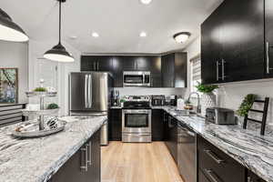 Kitchen featuring dark cabinetry, light stone countertops, appliances with stainless steel finishes, light wood finished floors, and recessed lighting
