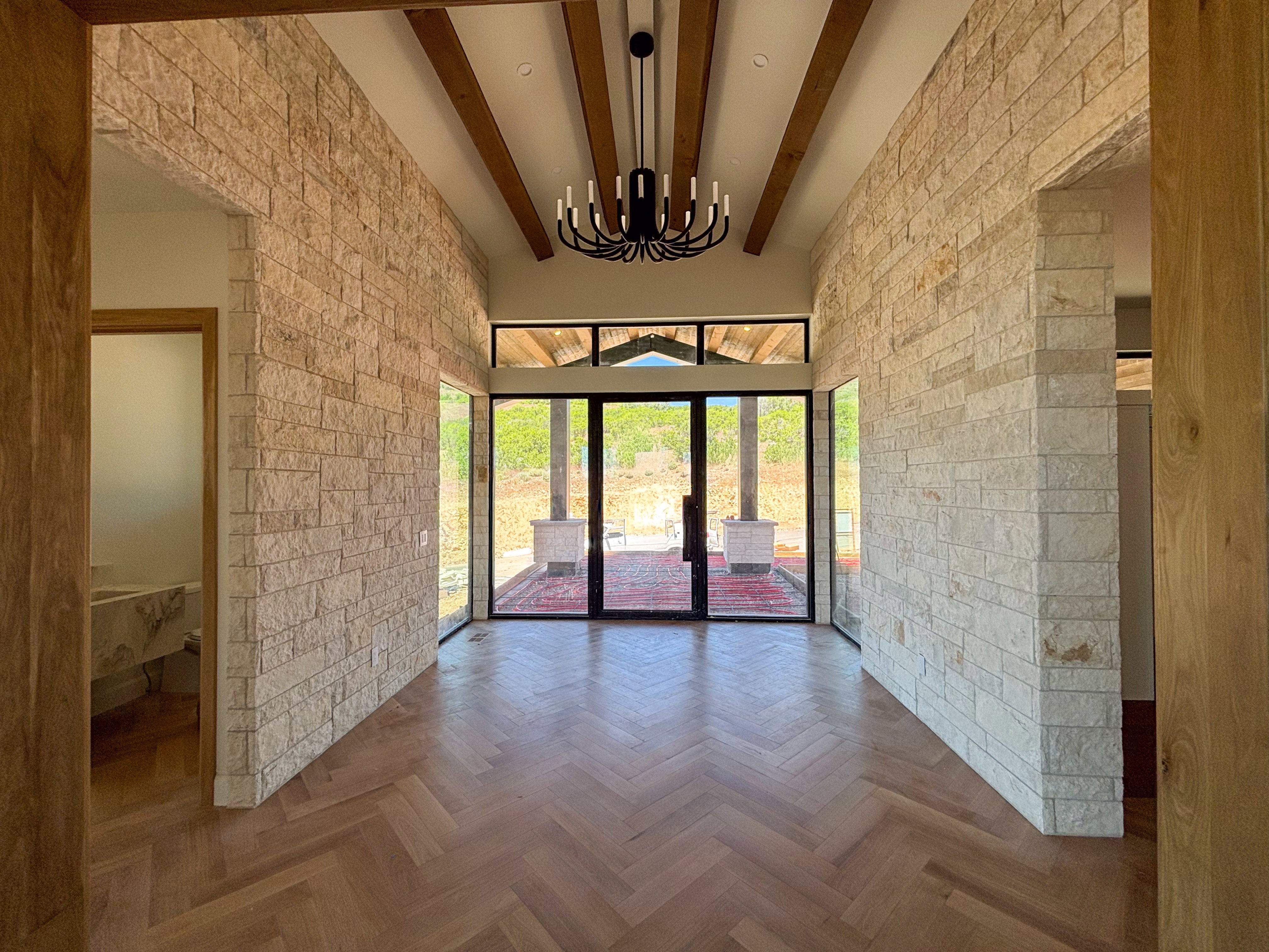 Entryway featuring herringbone wood-finish flooring, stone-clad walls, exposed wood ceiling beams, a black candelabra chandelier, and floor-to-ceiling windows