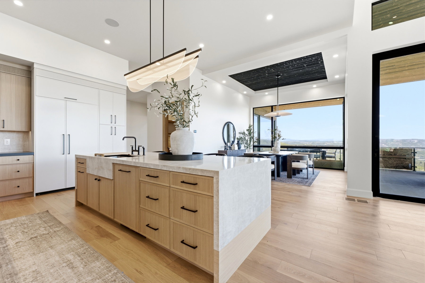 Open-concept kitchen and dining area featuring a spacious island with a farmhouse sink, light wood-finish flooring, full-height white cabinetry, and extensive window walls offering distant landscape scenery