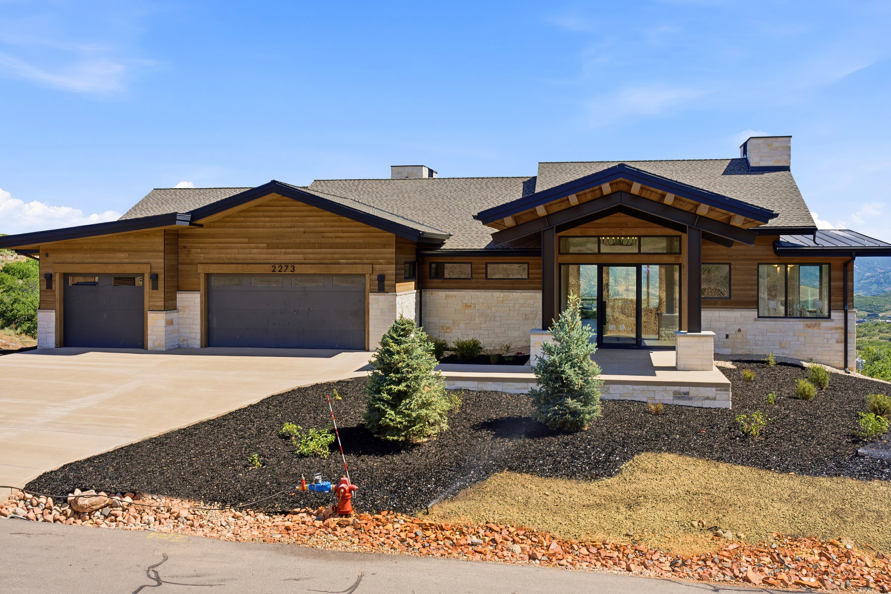 Contemporary exterior showcasing natural wood siding, stone accents, a deep-set covered entry, expansive glass doors, and a multi-car garage