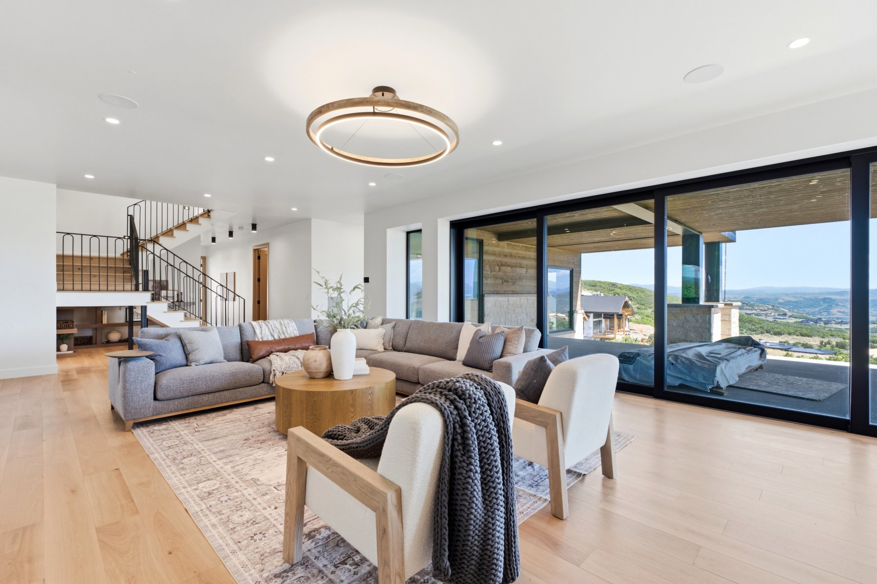 Spacious living area featuring light wood-finish flooring, a contemporary chandelier, recessed lighting, and a prominent staircase with black metal railings