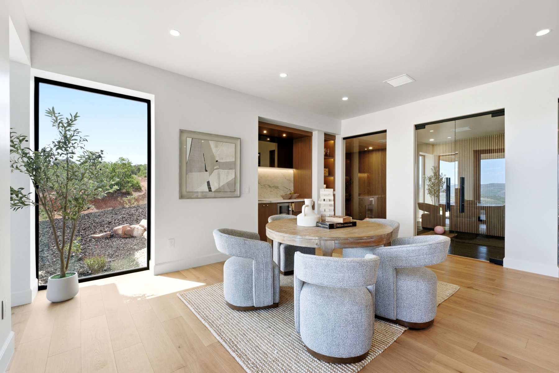 Spacious interior featuring light wood-finish flooring, a large floor-to-ceiling window, a built-in wet bar with shelving, and two glass-paneled entryways with black frames