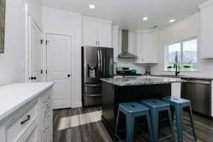 Kitchen with appliances with stainless steel finishes, light stone countertops, wall chimney exhaust hood, a breakfast bar, and dark wood-type flooring