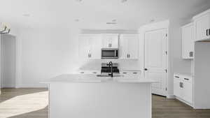 Kitchen with dark wood-style flooring, light stone countertops, stainless steel appliances, white cabinetry, and an island with sink