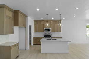 Kitchen featuring stainless steel appliances, light wood finished floors, pendant lighting, a center island with sink, and light stone countertops