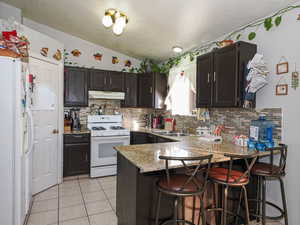 Kitchen with white appliances, dark brown cabinets, a breakfast bar, light tile patterned flooring, and light stone counters
