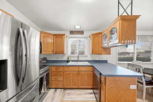 Kitchen featuring appliances with stainless steel finishes, a peninsula, dark countertops, brown cabinets, and glass insert cabinets