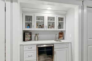 Indoor wet bar with white cabinetry, wine cooler, glass insert cabinets, and light stone counters