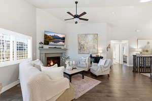 Living room with dark wood-type flooring, a warm lit fireplace, a ceiling fan, and high vaulted ceiling