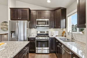 Kitchen featuring stainless steel appliances, light stone counters, dark brown cabinetry, vaulted ceiling, and decorative backsplash