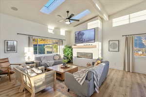 Living area with a glass covered fireplace, ceiling fan, light wood finished floors, high vaulted ceiling, and a skylight